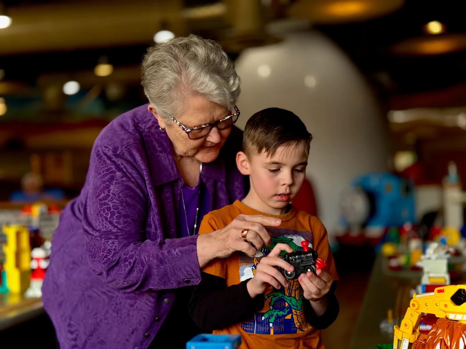 An older volunteer in a purple jacket leaning in beside a young boy at the museum's wooden train table, the two of them working on a toy engine together.