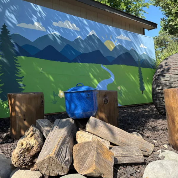 Cookware on logs set up for a fire in front of a mural of mountains
